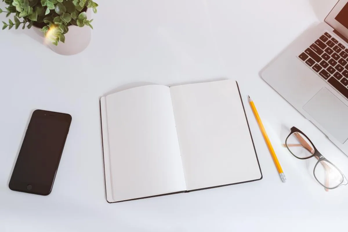 An open blank notebook on a white desk with a yellow pencil, a pair of glasses, a phone, a laptop corner and a green plant