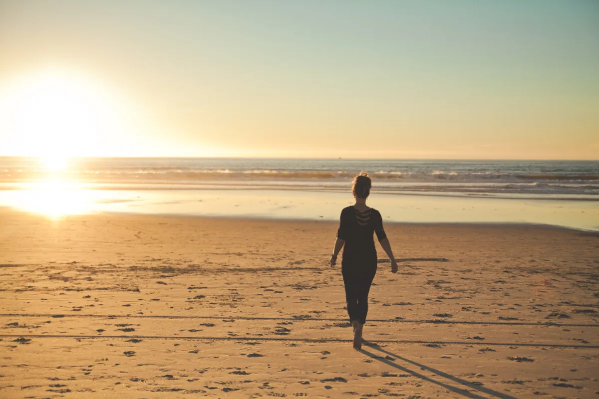A woman seen from behind walking barefoot across a sandy beach towards the sea in low evening sun