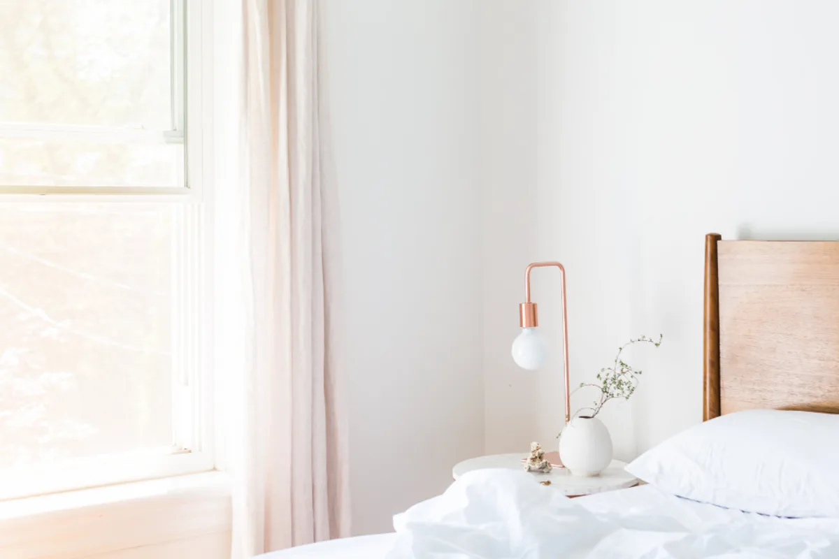 A bright bedroom corner with a tall window, a pale curtain, a copper lamp and a white vase on the bedside table
